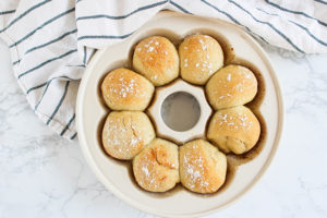 The Emile Henry Crown Baker with baked bread rolls sitting inside of it. These rest on a marble countertop and a navy striped tea towel