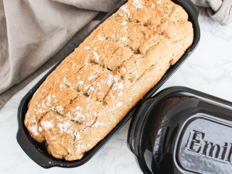 A loaf of baked bread cooling in the Emile Henry black glazed Italian Loaf Baker. Its lid is sitting to the side of the baker and all of it is resting on a light colored tea towel.
