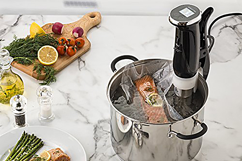 Salmon cooking in a sous vide device on a white marble countertop surrounded by other fresh ingredients