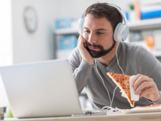Bearded man with headphones on with a laptop and a slice of pizza