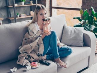 A blonde woman sits on a couch with a blanket wrapped around her eating a chocolate glazed donut