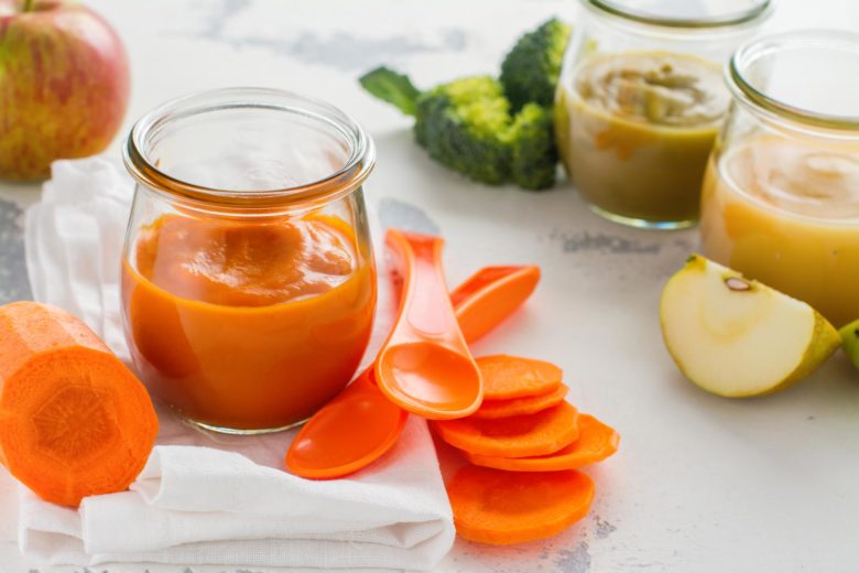 A jar of orange pureed baby food surrounded by small spoons, sliced carrots and other pureed veggies including broccoli