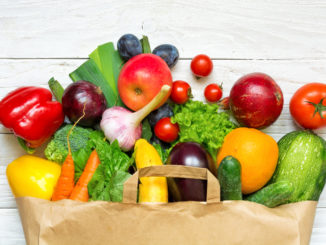 Full paper bag of different fruits and vegetables on a white wooden background.