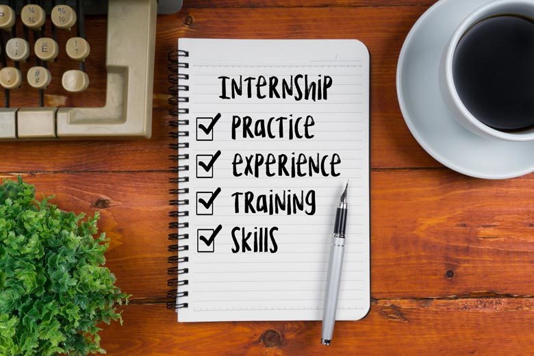 Notebook with pen on wooden desk background; notebook has title, "Internship," with checklist underneath with following items: practice, experience, training skills