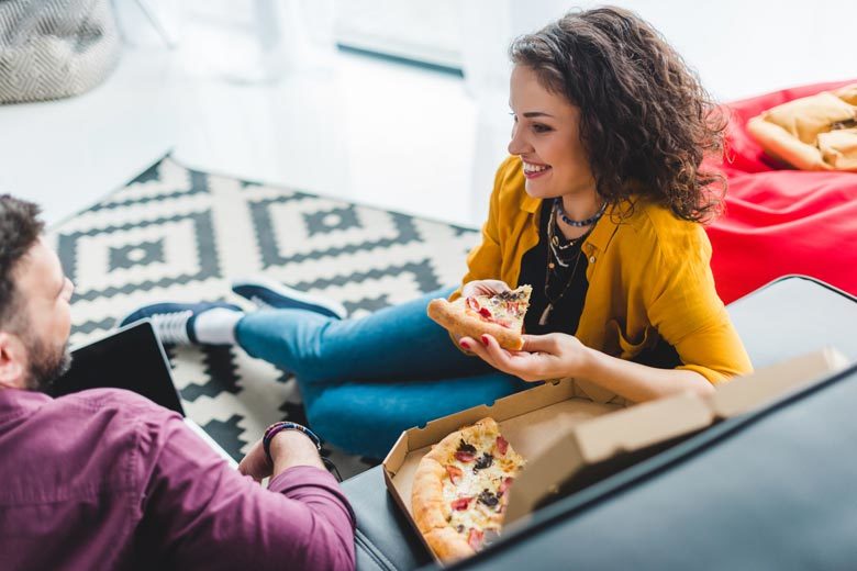 Two friends, a man and a woman, sitting on a couch eating pizza, talking and smiling