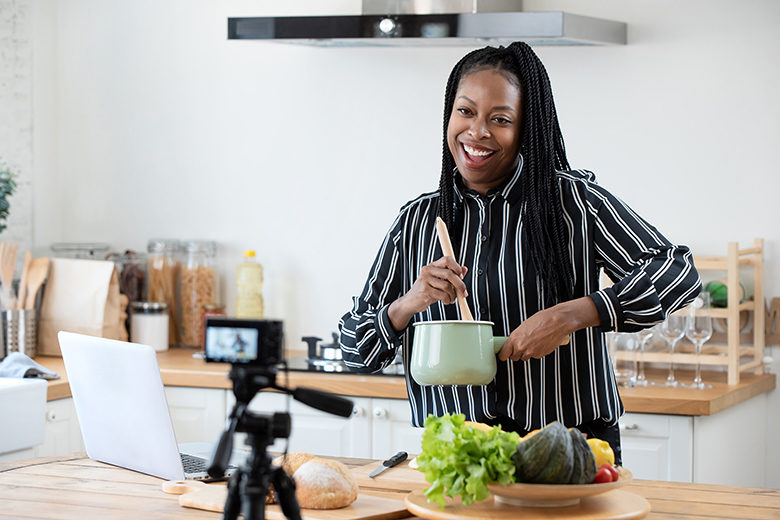 Happy woman vlogger broadcasting live video online while cooking food in kitchen at home